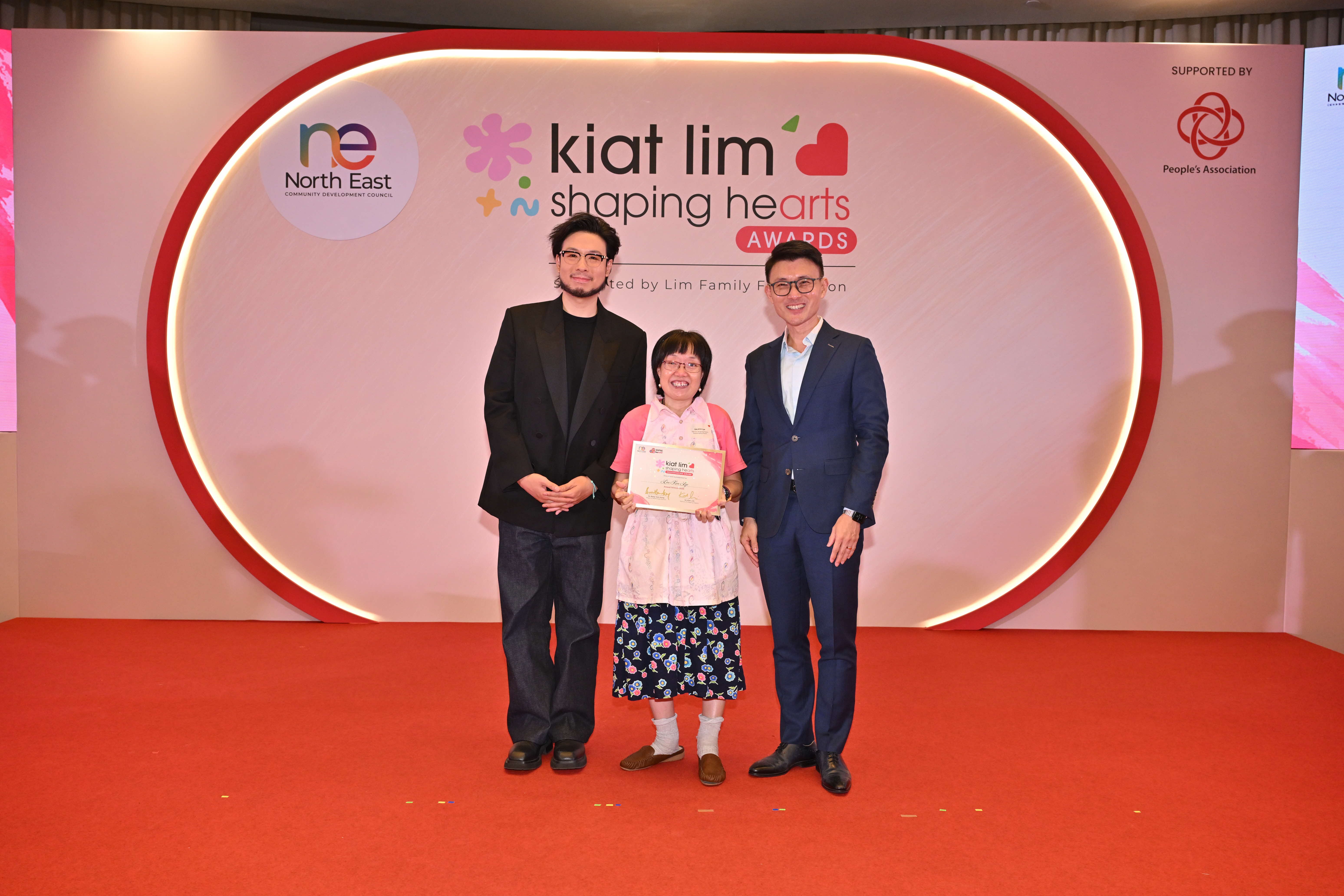 Award recipient standing on stage with the presenter, holding their certificate and posing for a group photo during the award ceremony, with a backdrop reading ‘Kiat Lim-Shaping Hearts Awards'.